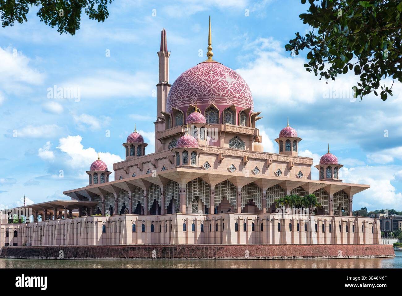 Putra Moschee, eine prächtige rosa Kuppelmoschee mit einem hohen Minarett, eingerahmt von Laub vor einem blauen bewölkten Himmel und Wasser in Putrajaya, Malaysia Stockfoto