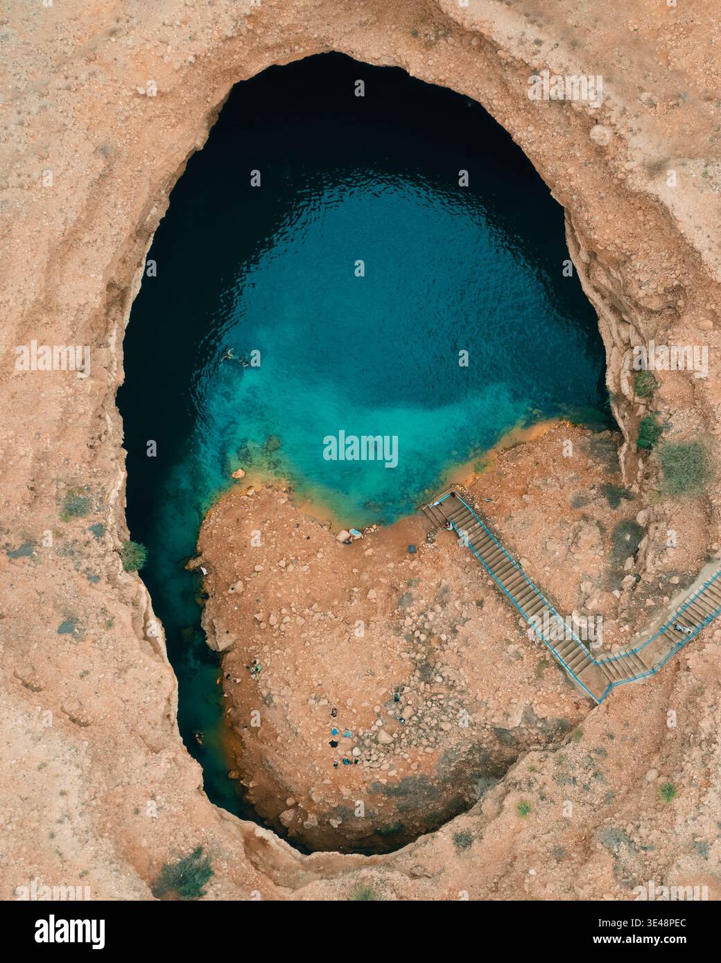 Blick aus der Vogelperspektive auf das Sink Hole mit türkisfarbenem Wasser, umgeben von zerklüfteten Wüstenklippen und einer Steintreppe in Dhofar Governorate, Oman. Stockfoto