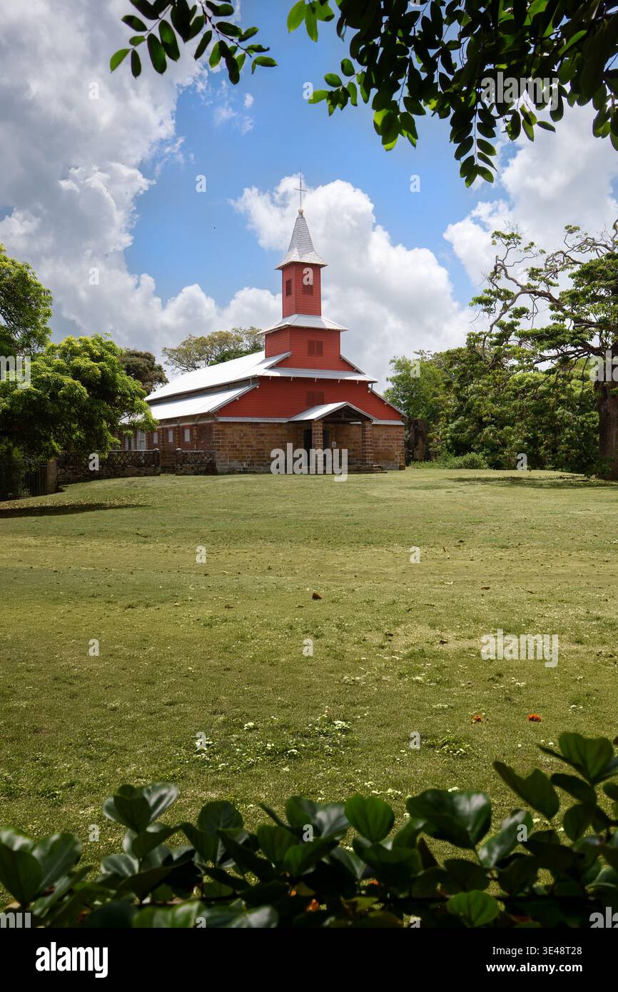 Blick auf die historische rot-weiße Kirche mit einem Steinfuß umgeben von üppigen grünen Bäumen und einem grasbewachsenen Feld unter einem bewölkten Himmel in Iles du Salut, Guy Stockfoto
