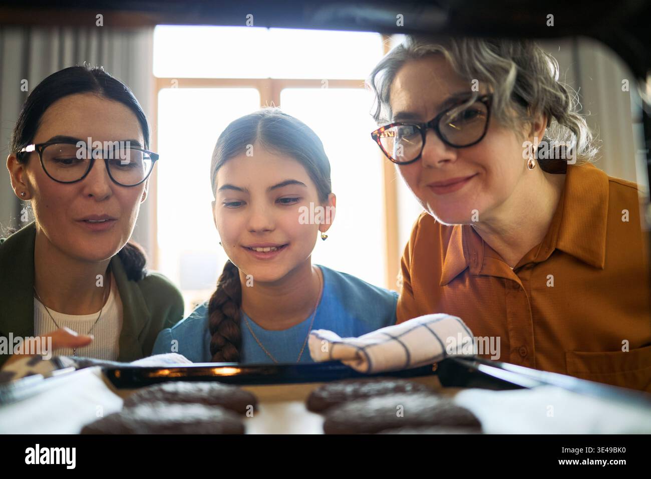 Frauen mittleren Alters mit Mädchen, die Kekse im Ofen checken, zu Hause zusammen backen und Familienaktivitäten teilen. Nützlich für Eltern, Generationen, Hausmannskost, Lifestyle-Marketing Stockfoto