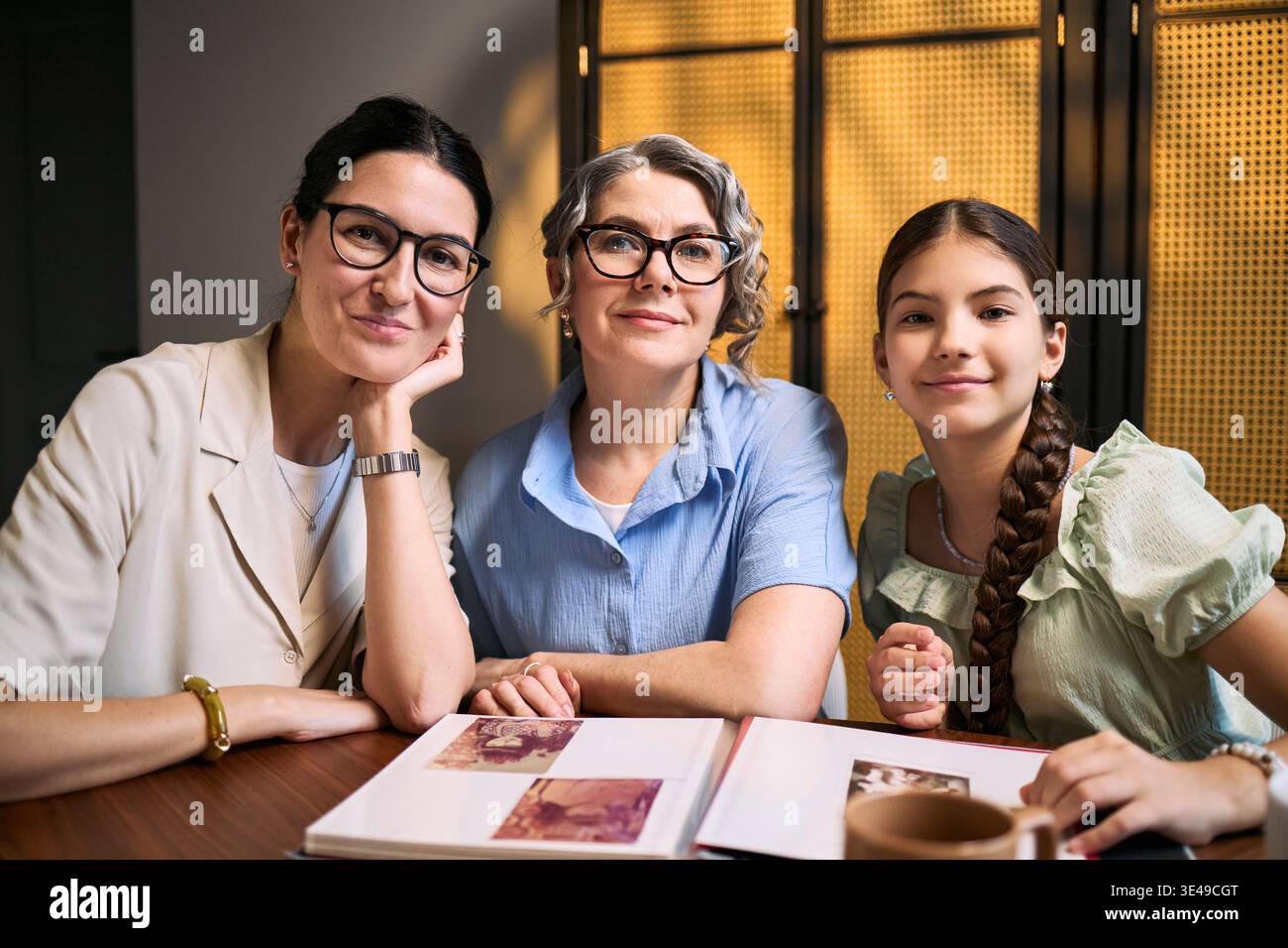 Porträt von Frauen aus mehreren Generationen, die ein Familienfoto-Album ansehen und die Kamera betrachten und dabei Erbe und Bindung darstellen. Nützlich für Familiendienstleistungen, Genealogie, Versicherungsmarketing Stockfoto