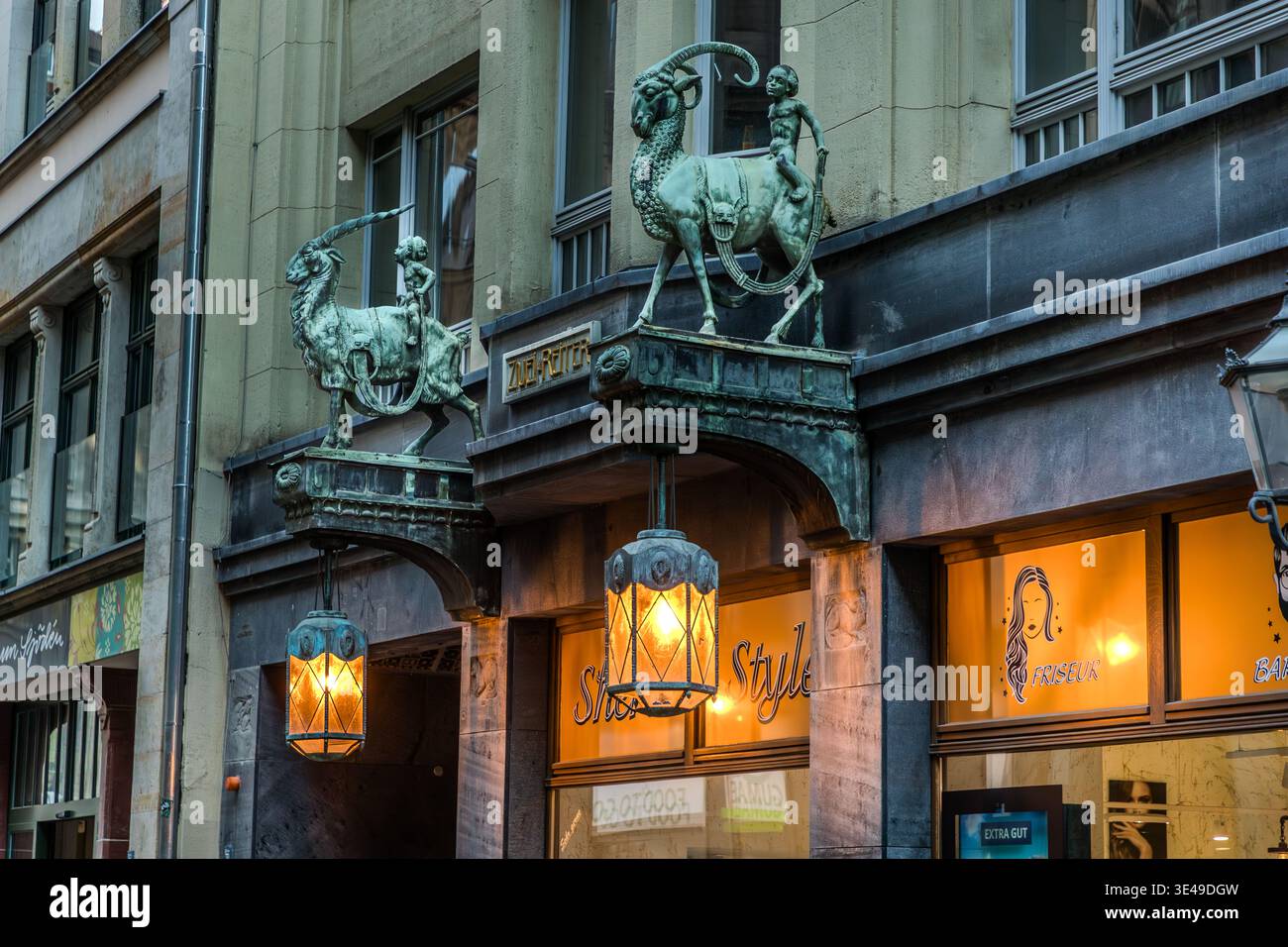 Zwei patinierte Bronzeskulpturen von Jungen auf Steinziegen flankieren den Eingang zu einem Jugendstilgebäude; beleuchtete Laternen hängen unter dem Eingangsbogen. Leipzig, Sachsen, Deutschland Stockfoto