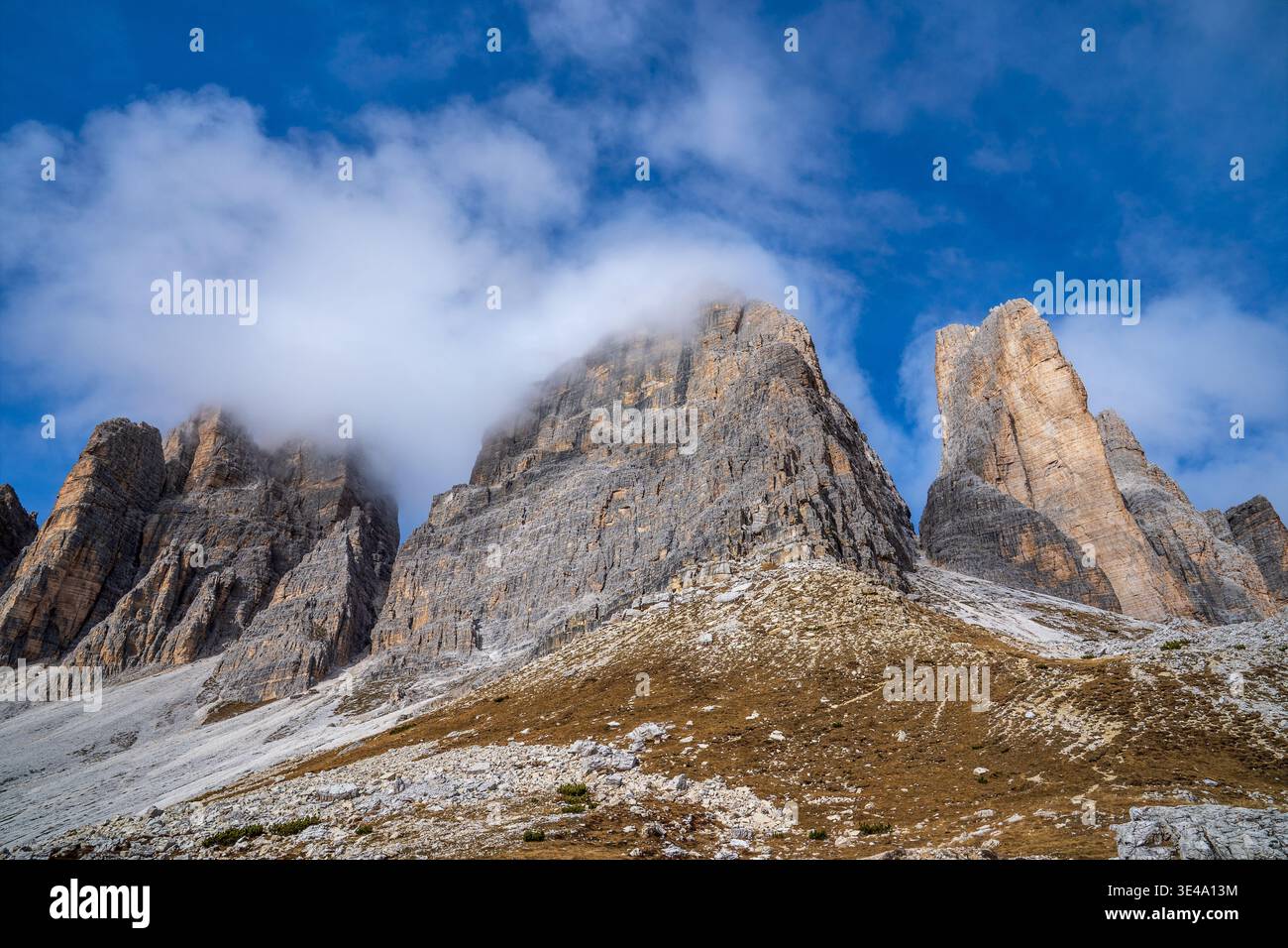 Tiefwinkelansicht der Gipfel der Tre Cime di Lavaredo. Die massiven Felswände schweben durch weiße Wolken und blauen Himmel, mit trockenem Gras und Schotterhängen im Fo Stockfoto