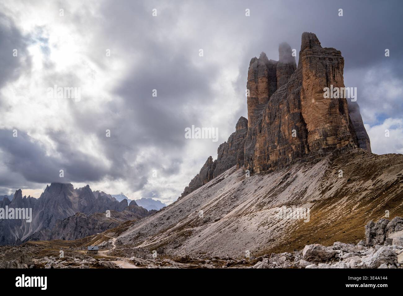 Seitenansicht der Tre Cime di Lavaredo mit Gipfeln, die in Nebel gehüllt sind. Ein unbefestigter Pfad und ein kleiner Unterschlupf darunter stehen im Kontrast zu den vertikalen Felswänden und dramatischen Stockfoto