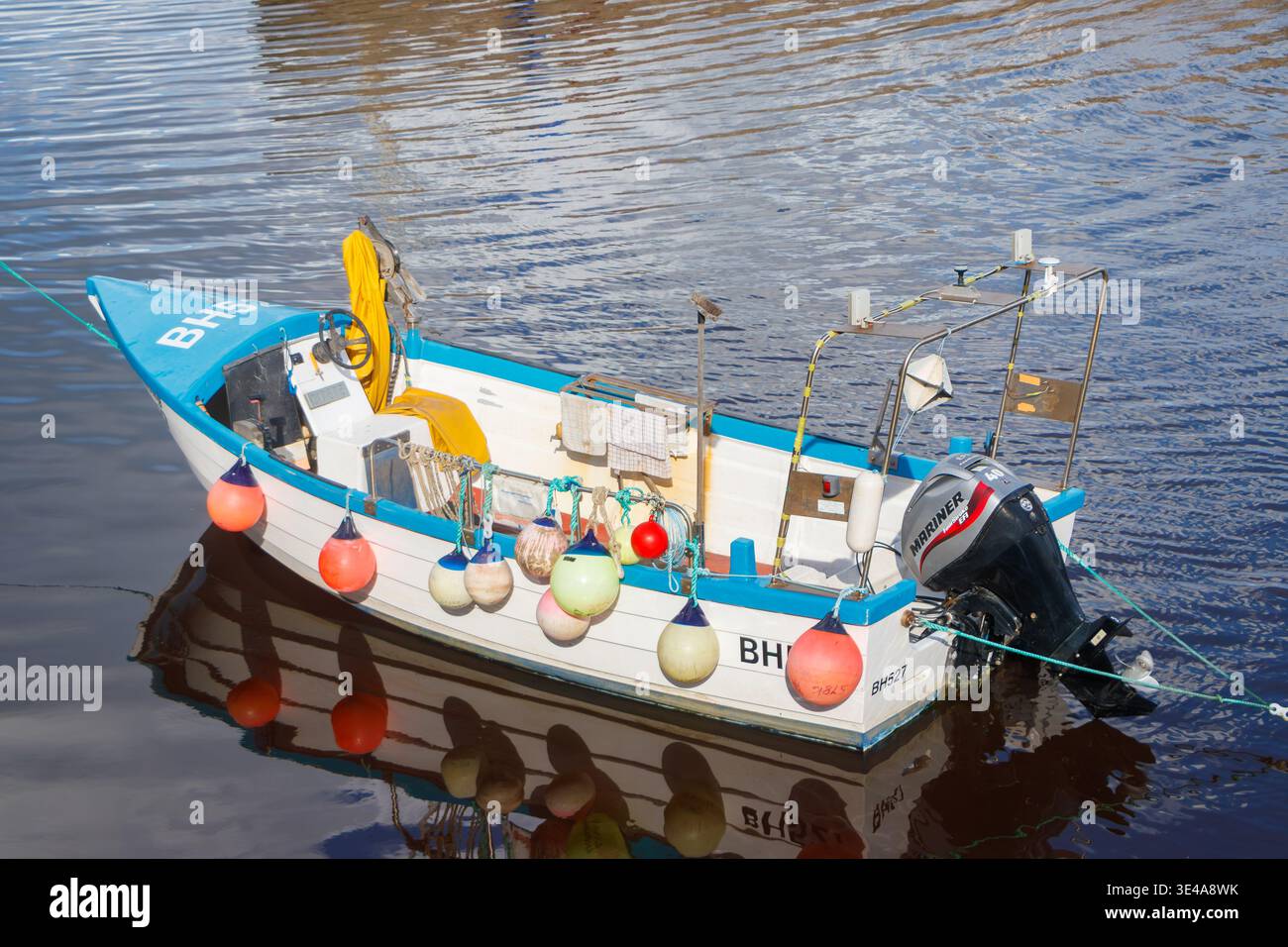 Kleines weißes und blaues offenes Fischerboot oder Coble mit einem Außenbordmotor des Mariners und farbenfrohen Bojen, die auf ruhigem Wasser verankert sind Stockfoto