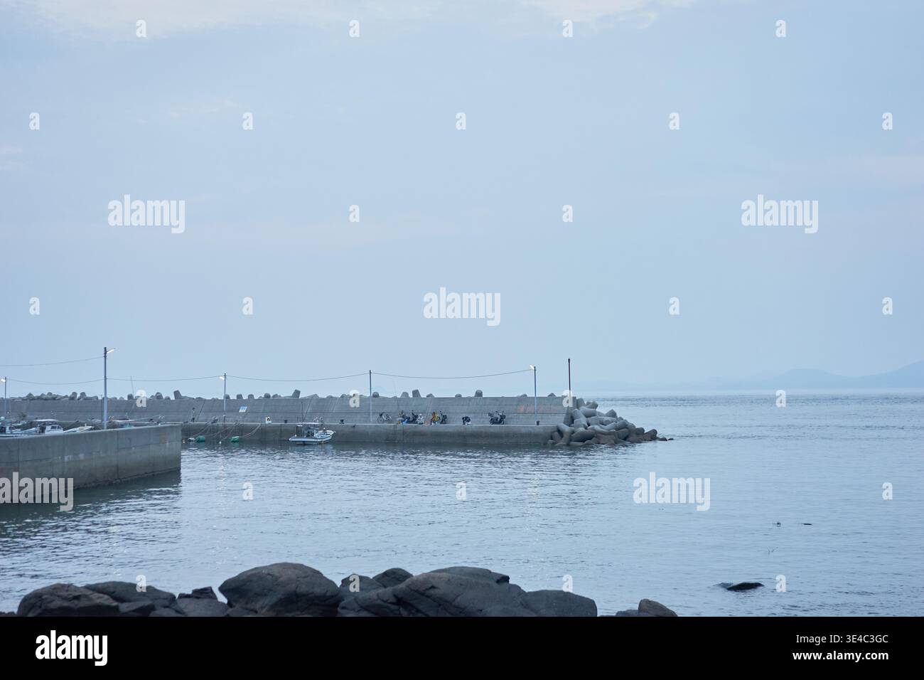 Ein ruhiger kleiner Hafen mit einem schwimmenden gelben Boot, einem roten Auto und Menschen, die einen entspannten Tag mit Angeln unter einem ruhigen, weichen, bedeckten Himmel genießen. Stockfoto