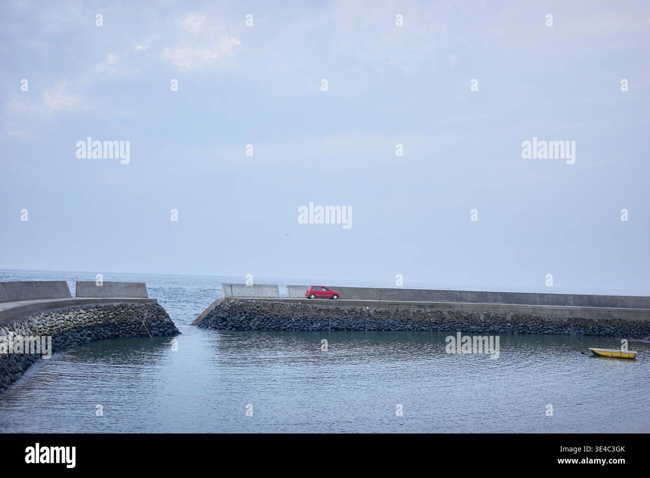 Ein ruhiger kleiner Hafen mit einem schwimmenden gelben Boot, einem roten Auto und Menschen, die einen entspannten Tag mit Angeln unter einem ruhigen, weichen, bedeckten Himmel genießen. Stockfoto