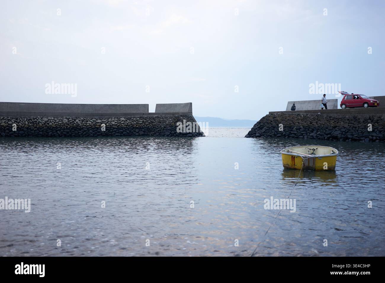 Ein ruhiger kleiner Hafen mit einem schwimmenden gelben Boot, einem roten Auto und Menschen, die einen entspannten Tag mit Angeln unter einem ruhigen, weichen, bedeckten Himmel genießen. Stockfoto