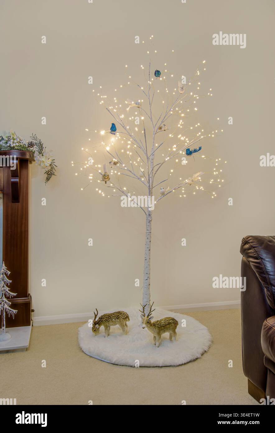 Beleuchteter weißer LED-Zweig mit festlichen Vögeln und künstlichen Hirschen in einem gemütlichen Innenraum Stockfoto