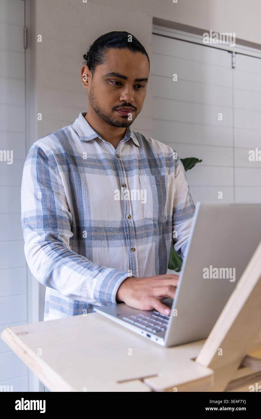 Asiatischer Mann tippt auf silbernem Laptop, steht am Schreibtisch und trägt kariertes Hemd im Büro mit Pflanze Stockfoto