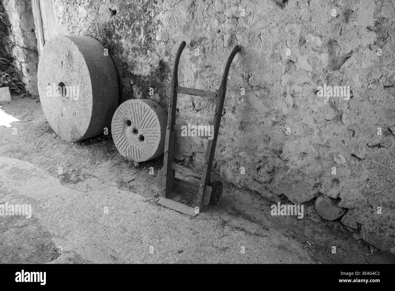Alte Mühlsteine und ein rustikaler Handwagen, der an einer Steinmauer in schwarz-weiß ruht, bayern, deutschland Stockfoto