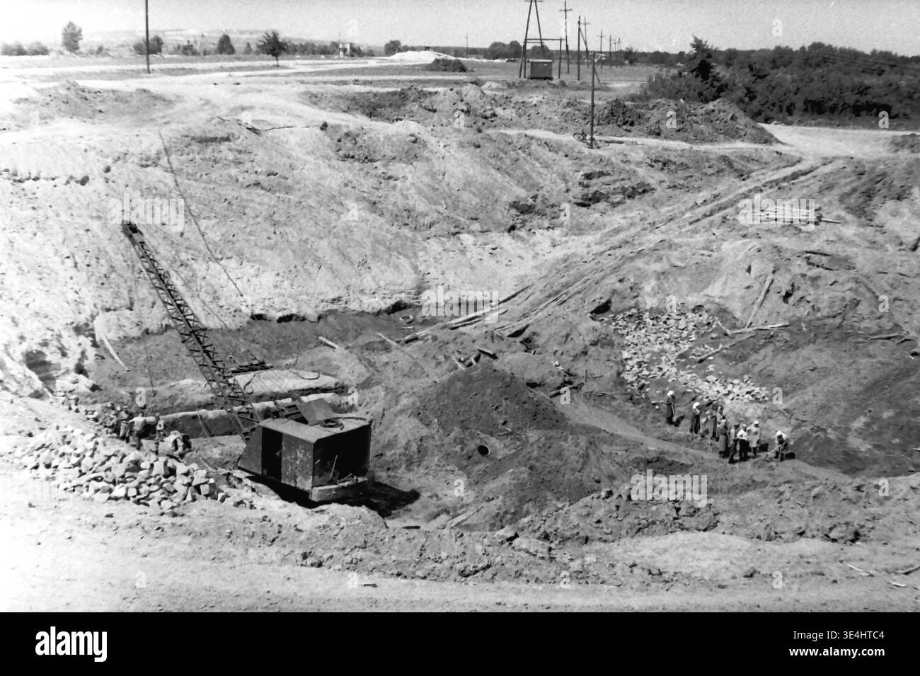 Nikolajewka, 1957. Slavyanskaja GRES-Kraftwerk Tiefgründungsgrube, sichtbar in einer faszinierenden Archivansicht. Ein schwerer mechanischer Schleppbagger befindet sich tief in dem riesigen Krater. Auf der rechten Seite ist eine engagierte Gruppe von Arbeitern, die zum Teil Kopftuch tragen, aktiv an intensiven manuellen Graben und Nivellieren des Bodens beteiligt. Dieses Vintage-Schwarzweiß-Foto stellt schwere Industriemaschinen und lebenswichtige menschliche Arbeit in einem wunderbaren Kontrast dar und symbolisiert das energische Vorkriegsleben und die schnelle industrielle Expansion im friedlichen Donbass. Stockfoto