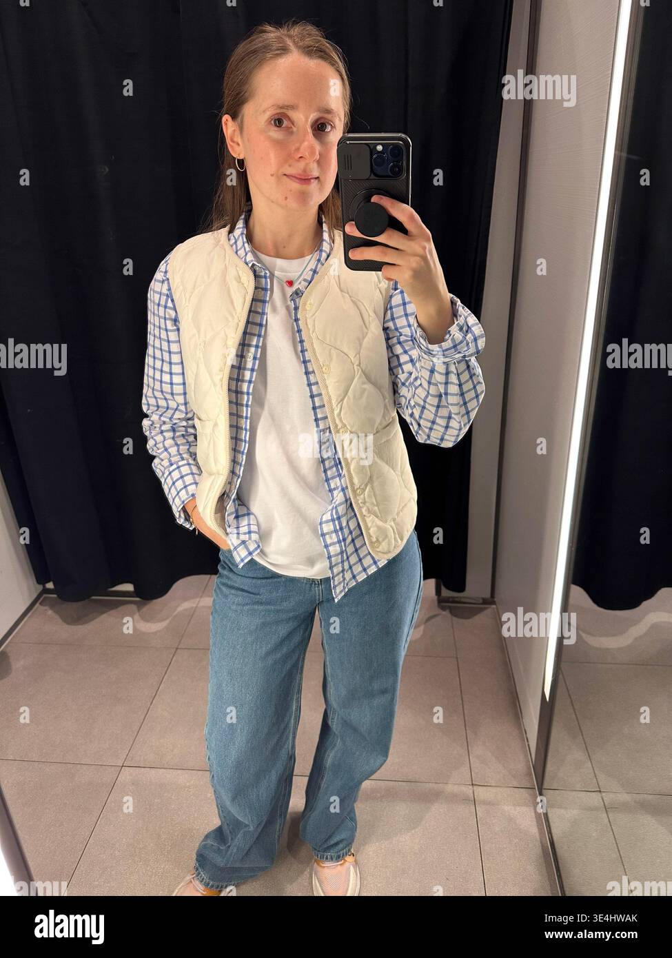 Junge Frau, die ein Spiegel-Selfie macht, während sie Kleidung in einer Garderobe anprobiert. Modernes Einkaufserlebnis, Modeauswahl Stockfoto