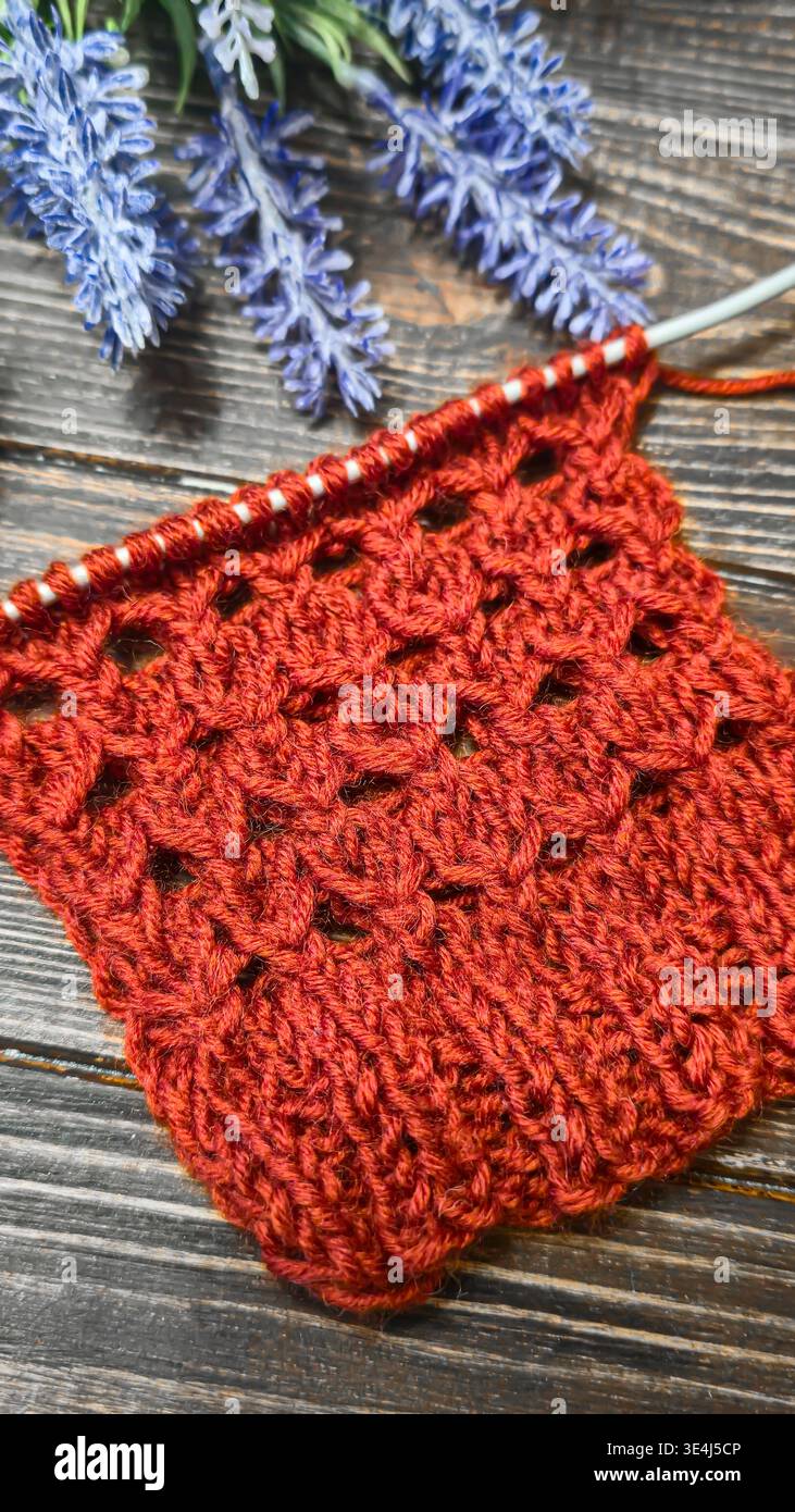 Ein strukturiertes, durchbrochenes Strickmuster aus farbigem Garn auf einer Holzoberfläche mit dekorativen Blumen. Nahaufnahme der Strickstruktur und des Strickmusters Stockfoto