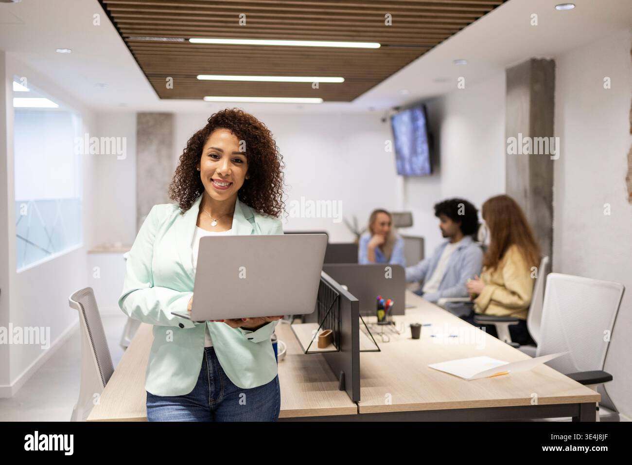 Selbstbewusster junger Profi, der einen Laptop in einem modernen Coworking-Büro hält, während Teamkollegen im Hintergrund zusammenarbeiten und Teamwork und Produ hervorheben Stockfoto