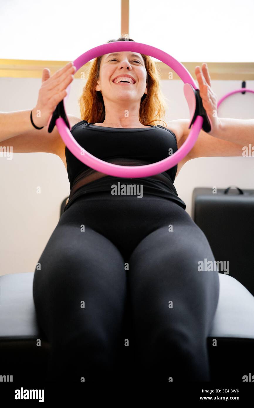Frau, die eine Pilates-Sitzung genießt, lächelt, während sie einen rosa Fitnessring benutzt. Die helle Atmosphäre und motivierende Bewegung fördern Positivität und Gesundheit. Stockfoto