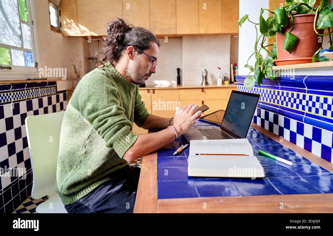 Ein junger hispanischer Mann sitzt an einem Tisch in einer Küche und benutzt seinen Laptop zur Arbeit. Er macht sich Notizen und konzentriert sich auf seine Aufgaben am Nachmittag. Pl Stockfoto