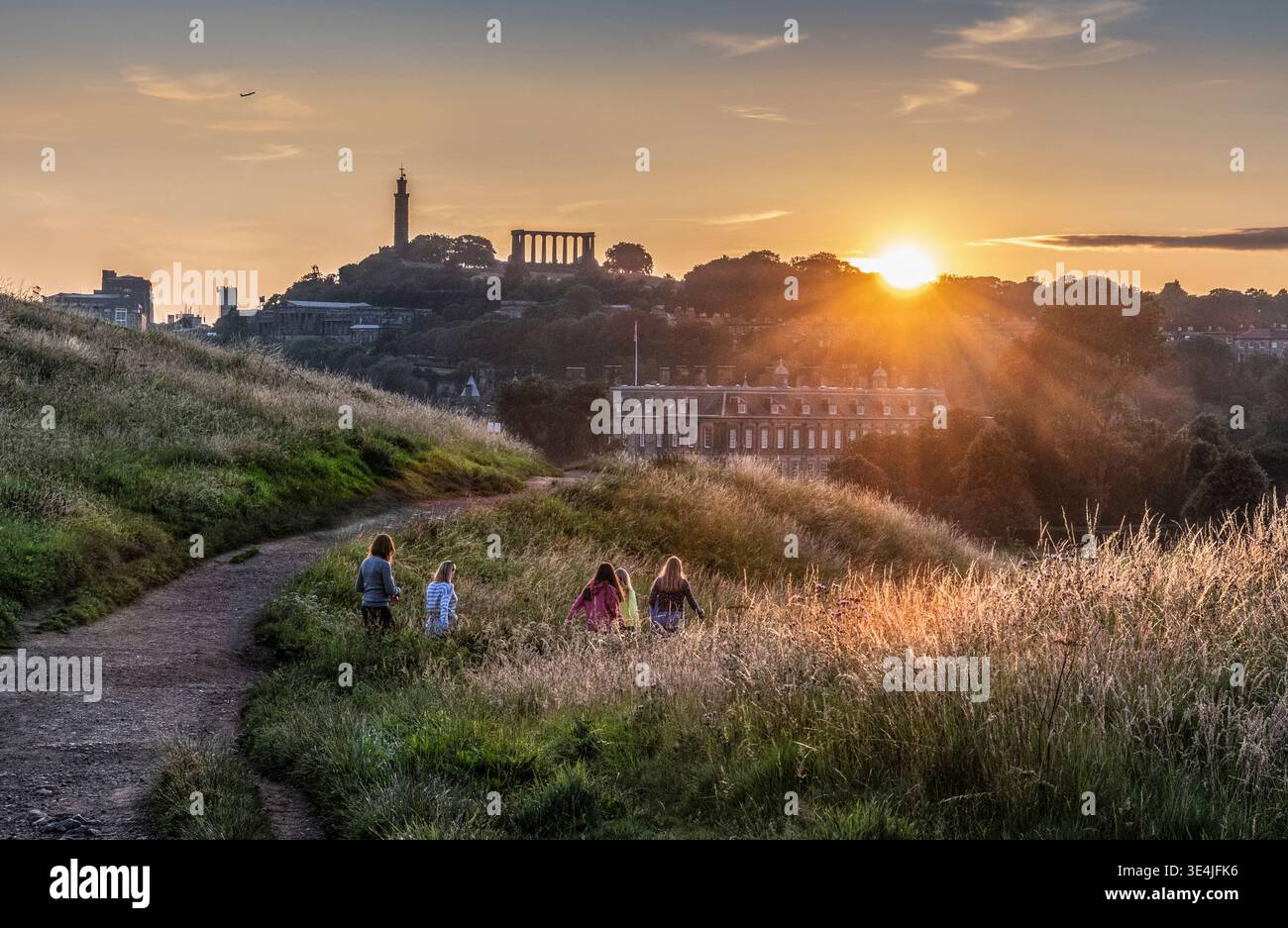 Eine Gruppe von Freunden, die bei Sonnenuntergang auf dem Calton Hill einen grasbewachsenen Hügel hinunterschlendern, mit einer Silhouette vor der goldenen Skyline von Edinburgh und einem warmen Sonnenuntergang, evoki Stockfoto