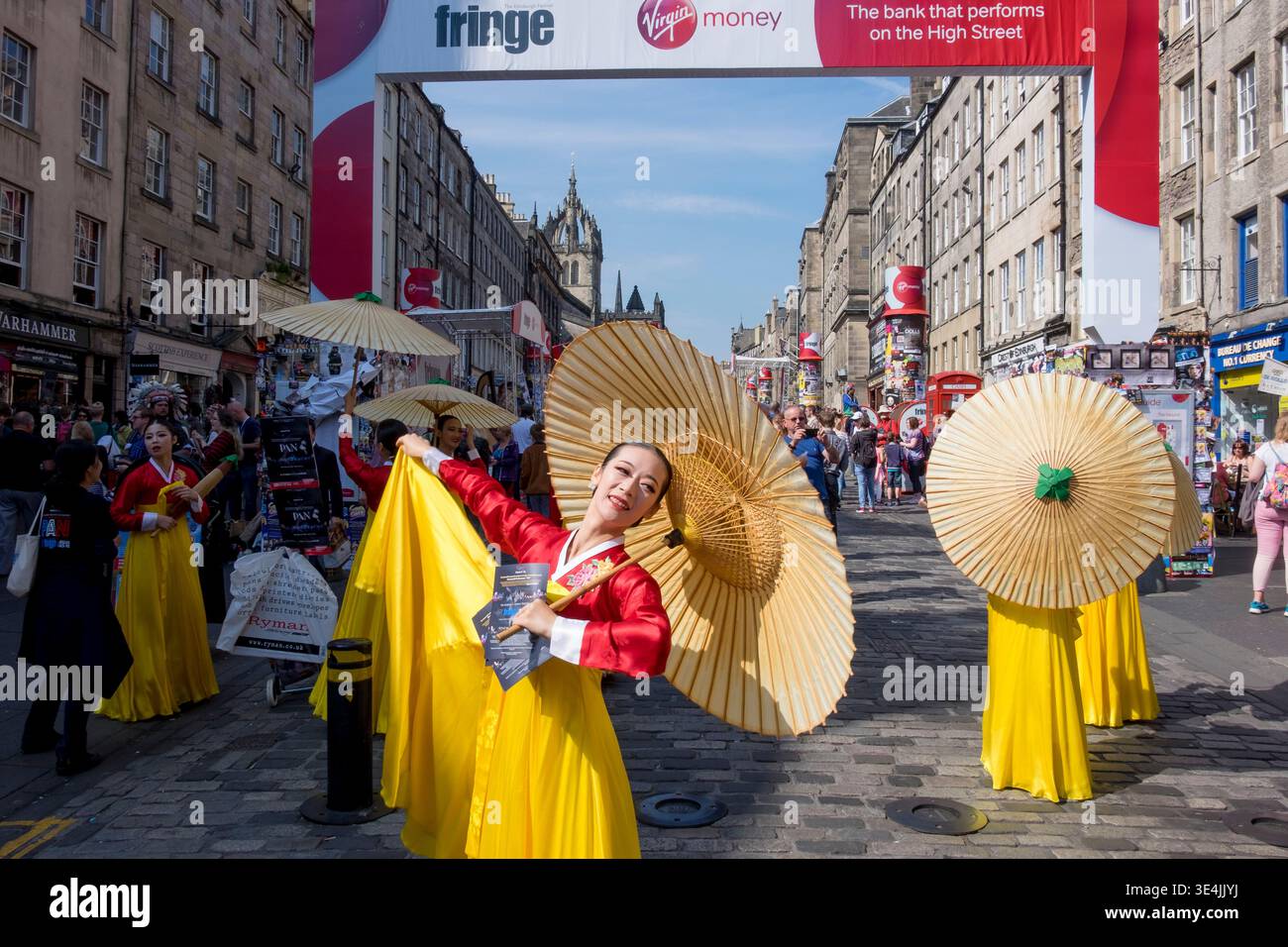 Farbenfrohe Straßenkünstler in gelben und roten Trachten tanzen während des Fringe Festivals mit Papierschirmen auf einer geschäftigen Edinburgh High Street Stockfoto