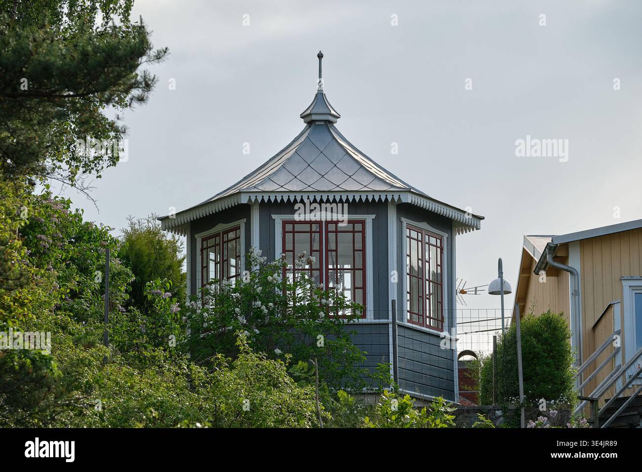 Ein kleines, weißes Gebäude mit grünem und rotem Dach. Das Dach ist schräg und hat eine spitze Oberseite. Das Gebäude ist von Bäumen und Büschen umgeben. N Stockfoto
