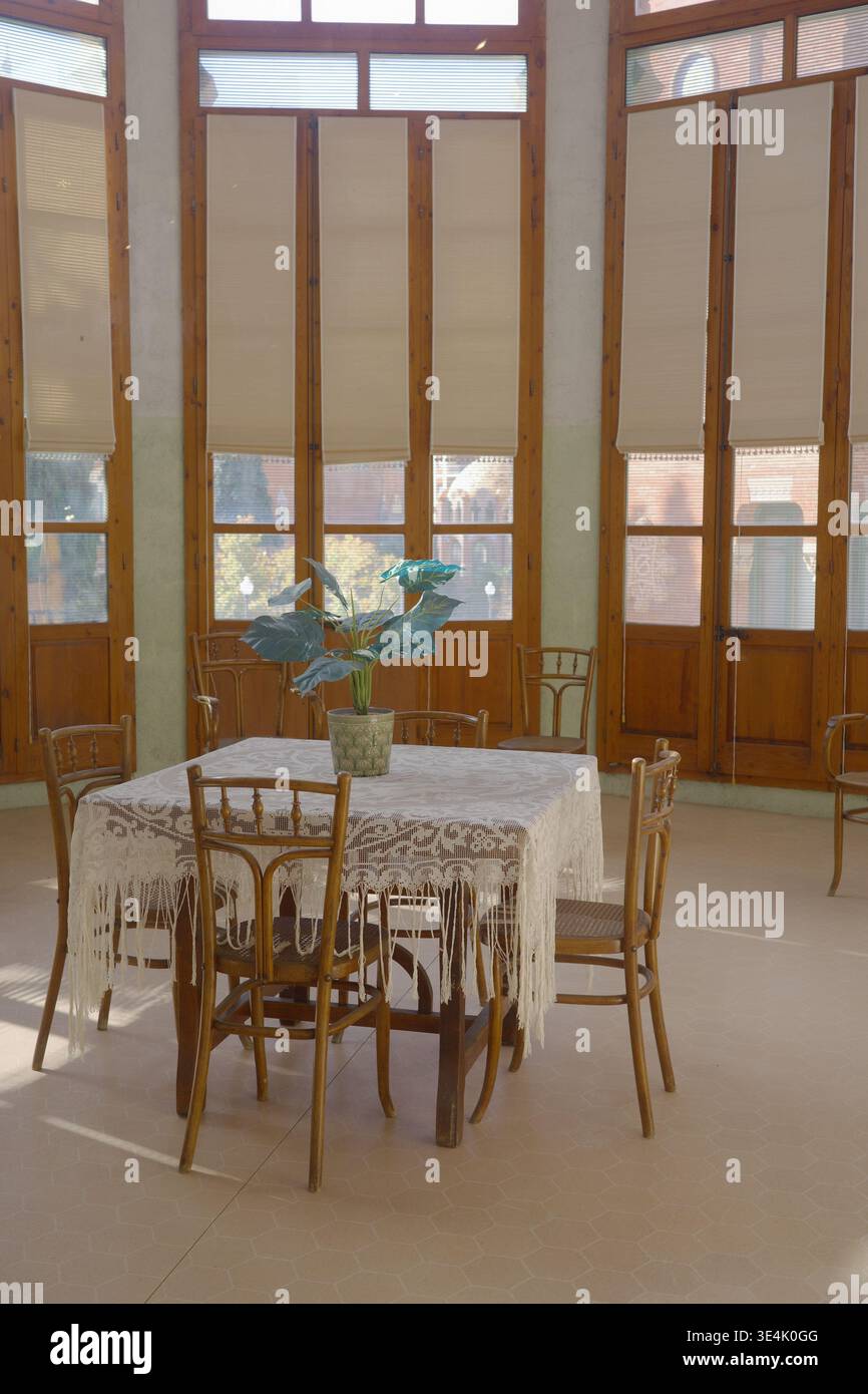 Sonnendurchfluteter Raum mit Thonet-Bugholzstühlen und Spitzentisch neben hohen Holzfenstern im Recinte Modernista de Sant Pau, Barcelona Stockfoto