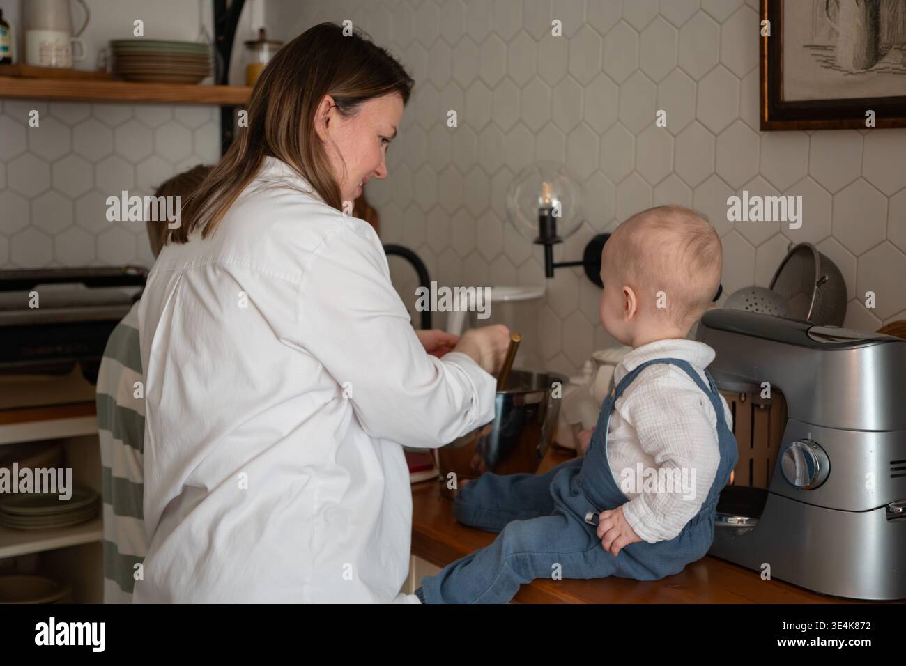 Die Mutter bereitet Zutaten zu, während das Baby während des Backens zu Hause auf der Küchenarbeitsplatte sitzt. Erziehung, Familienkochen, Kinderbeteiligung, häuslich Stockfoto