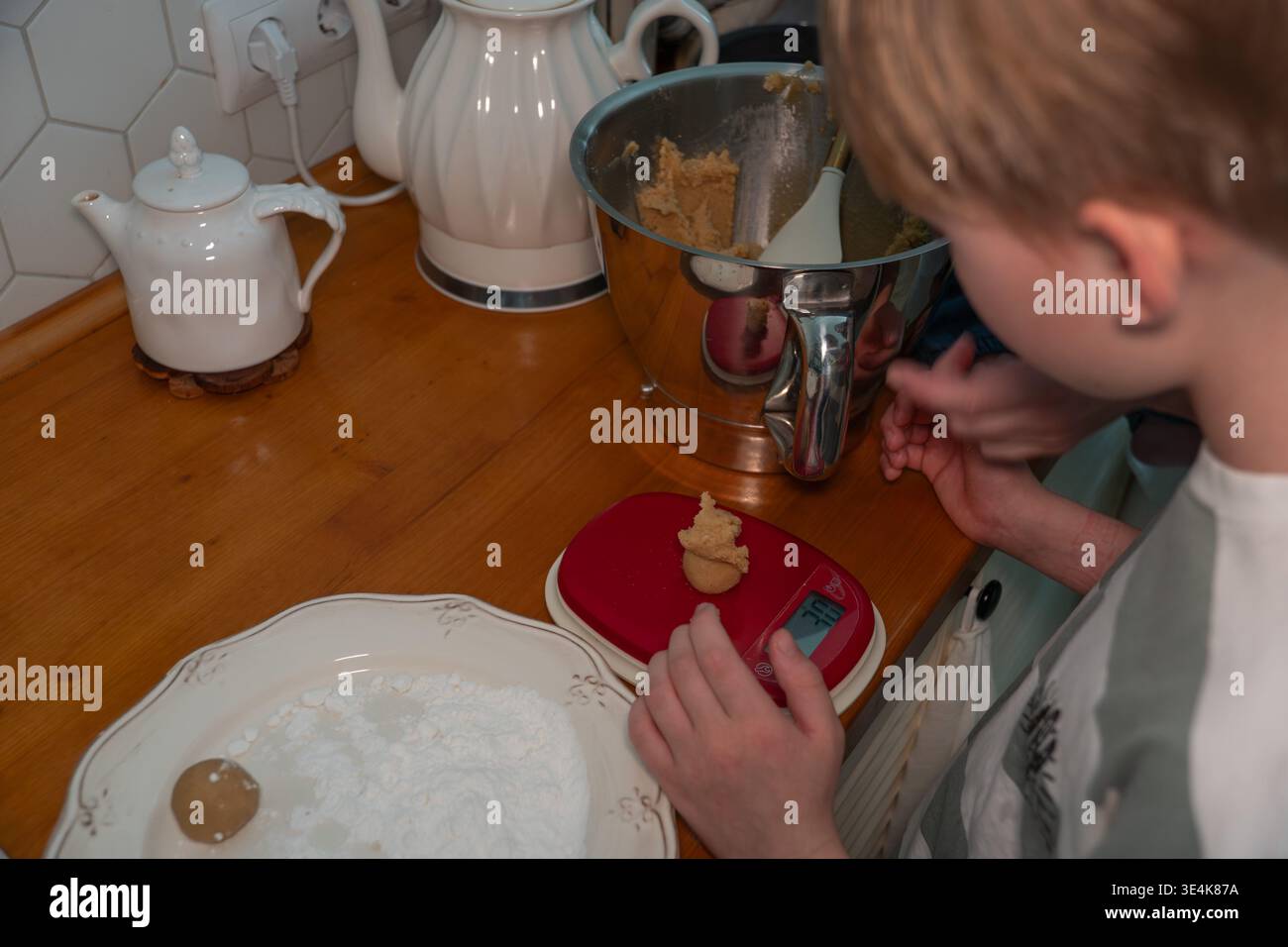 Das Kind wiegt Teigstücke auf der Küchenwaage während des Backprozesses zu Hause und erlernt Kochfähigkeiten in der Familienküche. Kinderbeteiligung, Familienkochen Stockfoto