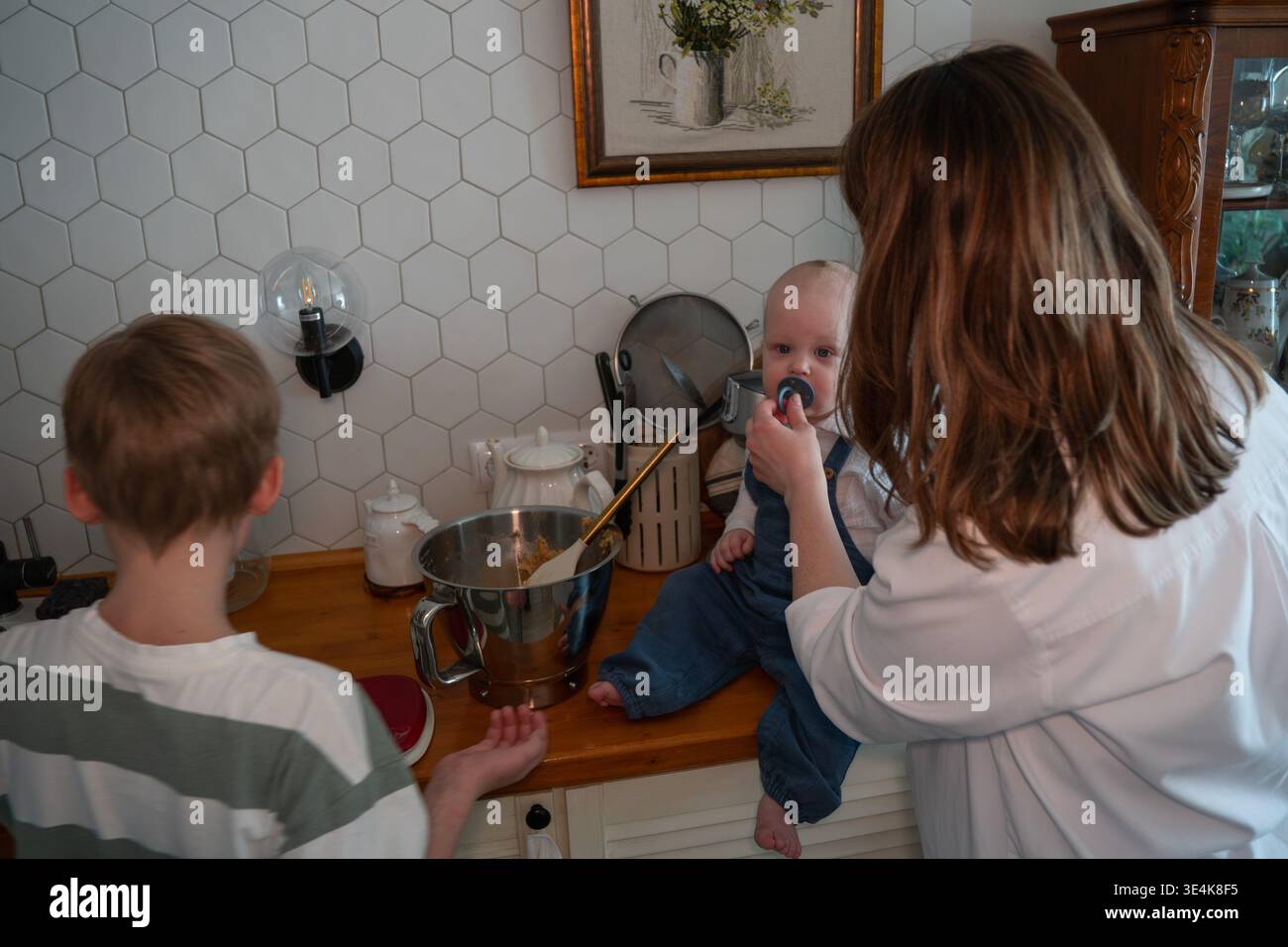 Die Mutter füttert das Baby, während die Familie während des Backens zu Hause Teig in der Küche zubereitet. Erziehung, Familienkochen, Kinderbeteiligung, häuslicher Lebensstil Stockfoto