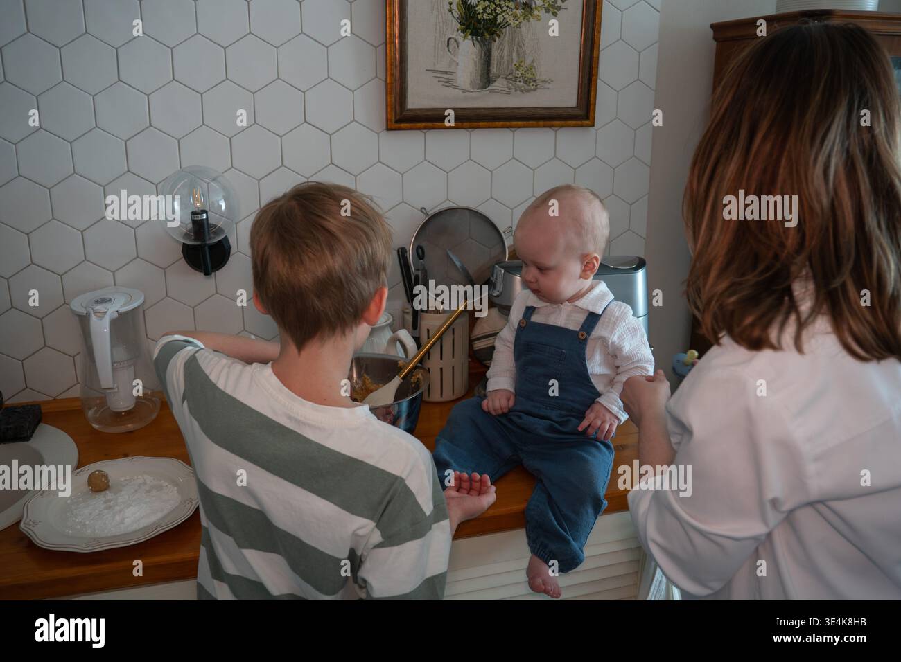 Familie backen zusammen in der Küche, Mutter und Kinder bereiten Teig zu, während das Baby auf der Arbeitsplatte sitzt. Hausmannskost, Elternschaft, Familienbindung Stockfoto