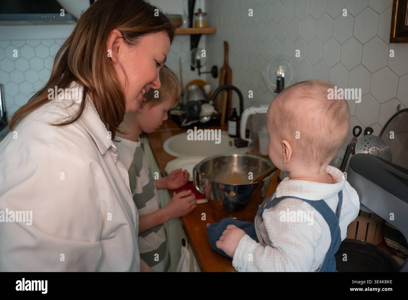 Die Mutter bereitet Zutaten zu, während das Baby während des Backens zu Hause auf der Küchenarbeitsplatte sitzt. Erziehung, Familienkochen, Kinderbeteiligung, häuslich Stockfoto