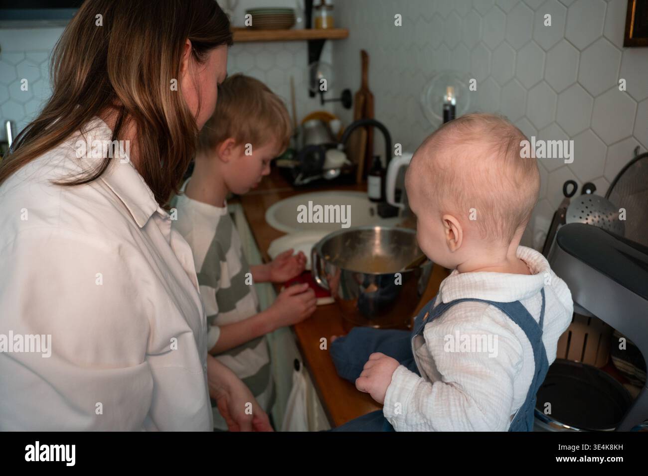 Die Mutter bereitet Zutaten zu, während das Baby während des Backens zu Hause auf der Küchenarbeitsplatte sitzt. Erziehung, Familienkochen, Kinderbeteiligung, häuslich Stockfoto