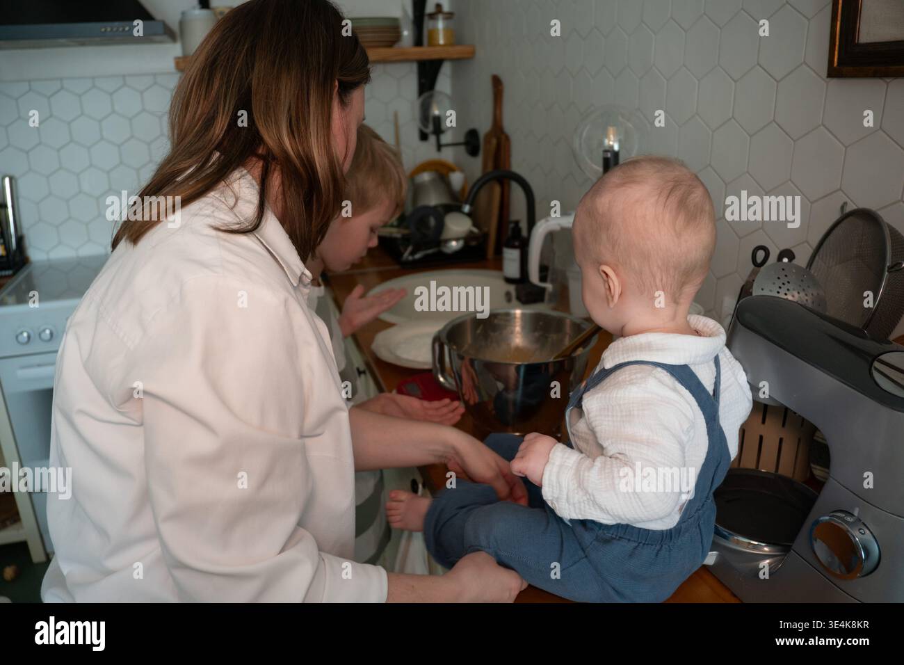 Die Mutter bereitet Zutaten zu, während das Baby während des Backens zu Hause auf der Küchenarbeitsplatte sitzt. Erziehung, Familienkochen, Kinderbeteiligung, häuslich Stockfoto