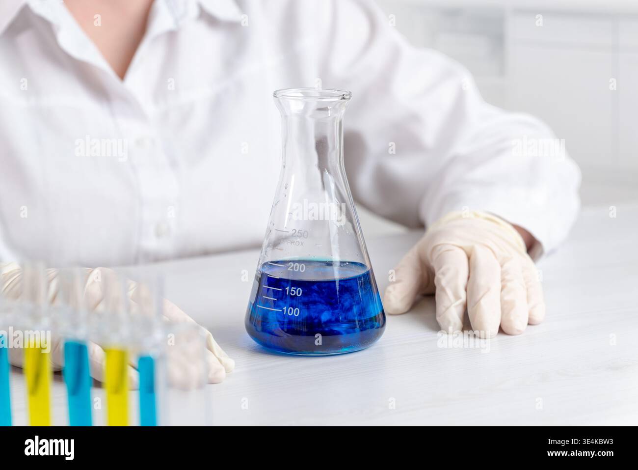 Laborkolben mit blauer chemischer Lösung und dunkler Reaktion im Inneren, Wissenschaftlerhände und Reagenzgläser im Hintergrund, wissenschaftliches Forschungskonzept Stockfoto