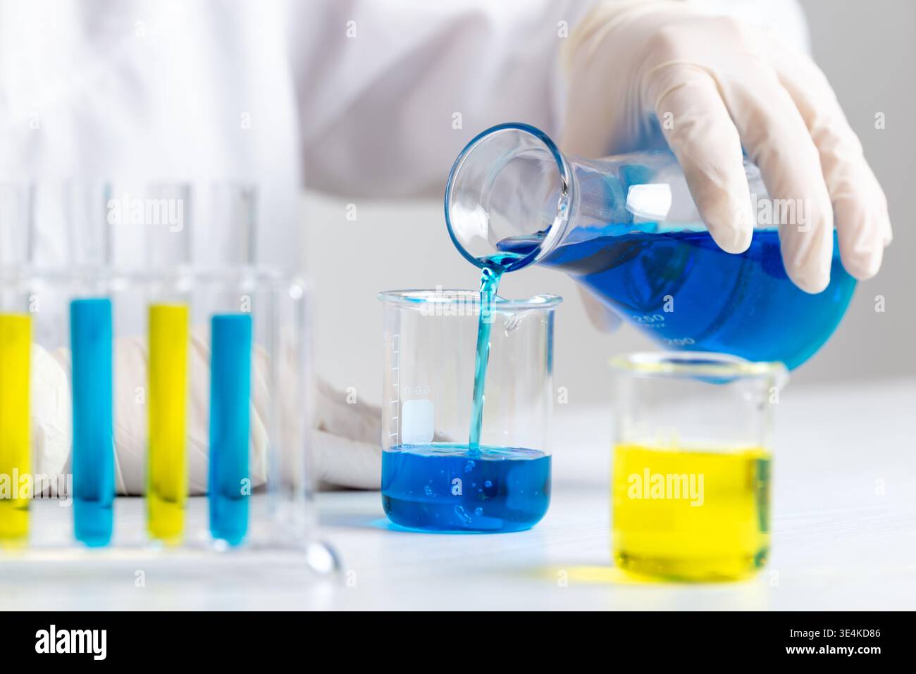 Wissenschaftler gießen blaue Flüssigkeit aus dem Kolben in Becher, Laborversuch und chemisches Mischen in kontrollierter Umgebung Stockfoto
