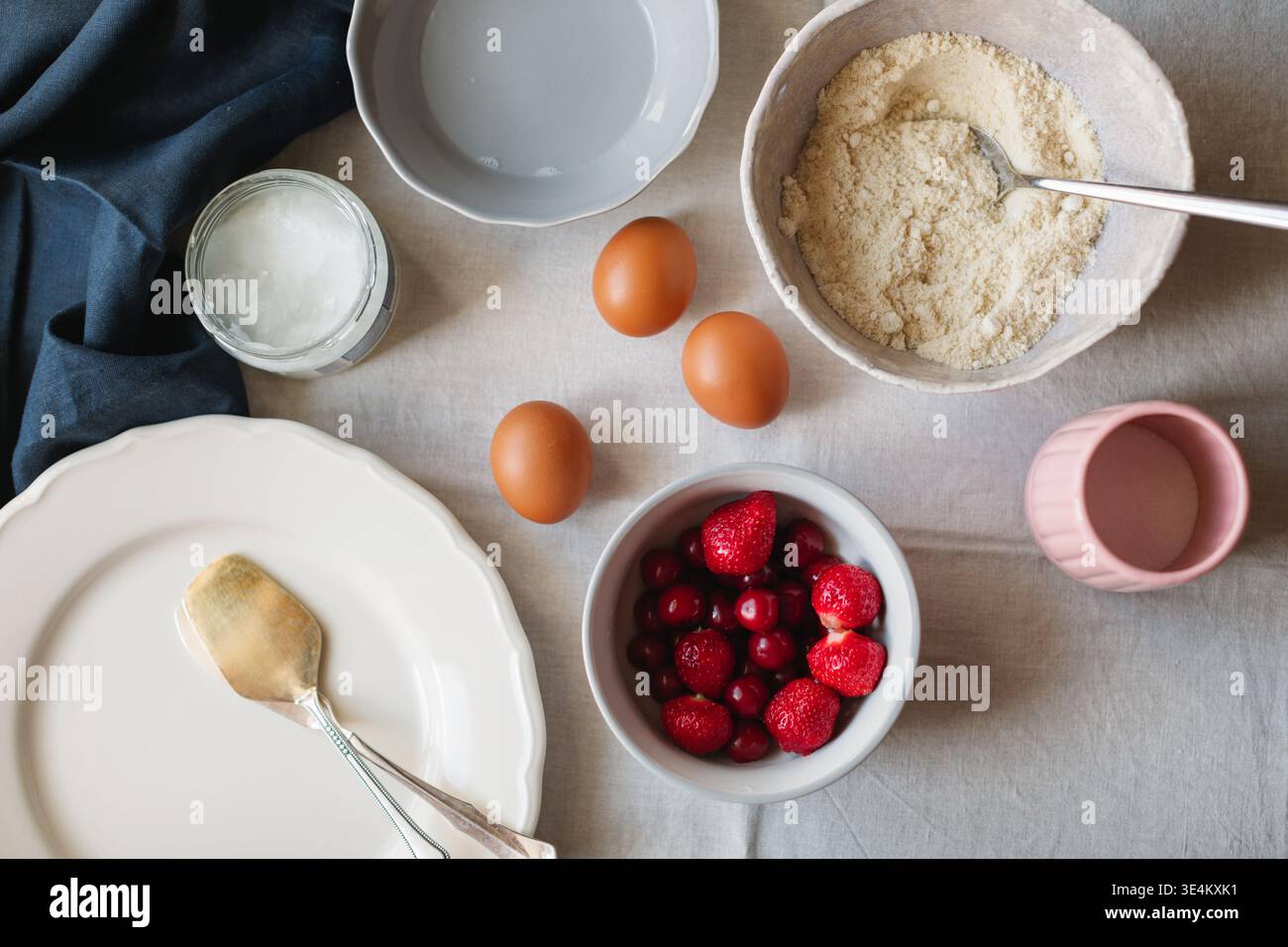 Blick von oben auf verschiedene Backzutaten für die Zubereitung eines Kuchens, einschließlich Eiern, Erdbeeren, Kirschen, Kokosöl und glutenfreiem Mandel- und Kokosmehl Stockfoto
