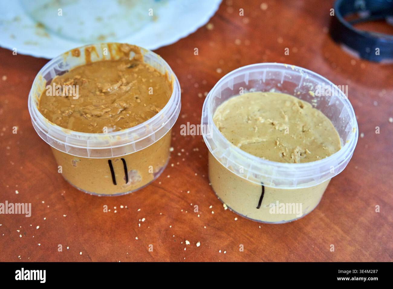 Zwei transparente Kunststoffbehälter, gefüllt mit dicker Nussbutter, sitzen auf einem rotbraunen Holztisch, der mit kleinen weißen Krümeln verstreut ist. Links Stockfoto