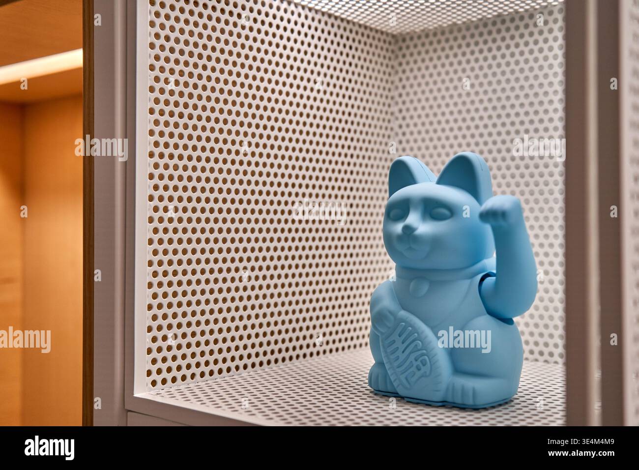 Eine hellblaue maneki-neko-Figur sitzt auf einem weißen perforierten Metallregal in einem modernen Schrank. Die japanische Glückskatzenstatue hat es Stockfoto