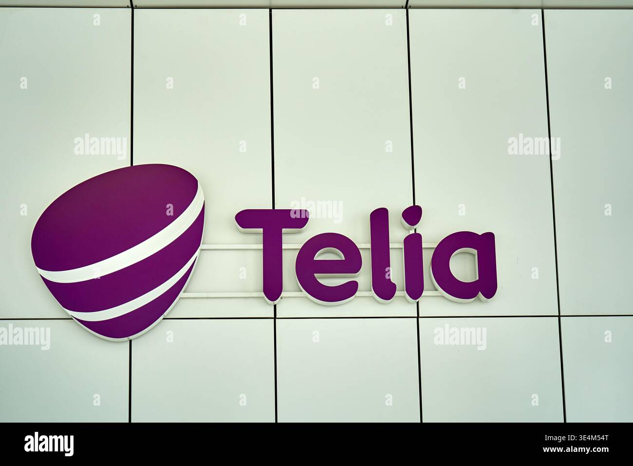 Das Bild zeigt das Firmenlogo von Telia, einem Telekommunikationsunternehmen, das auf einer hellen, gekachelten Wand montiert ist. Das Logo besteht aus einem Stockfoto
