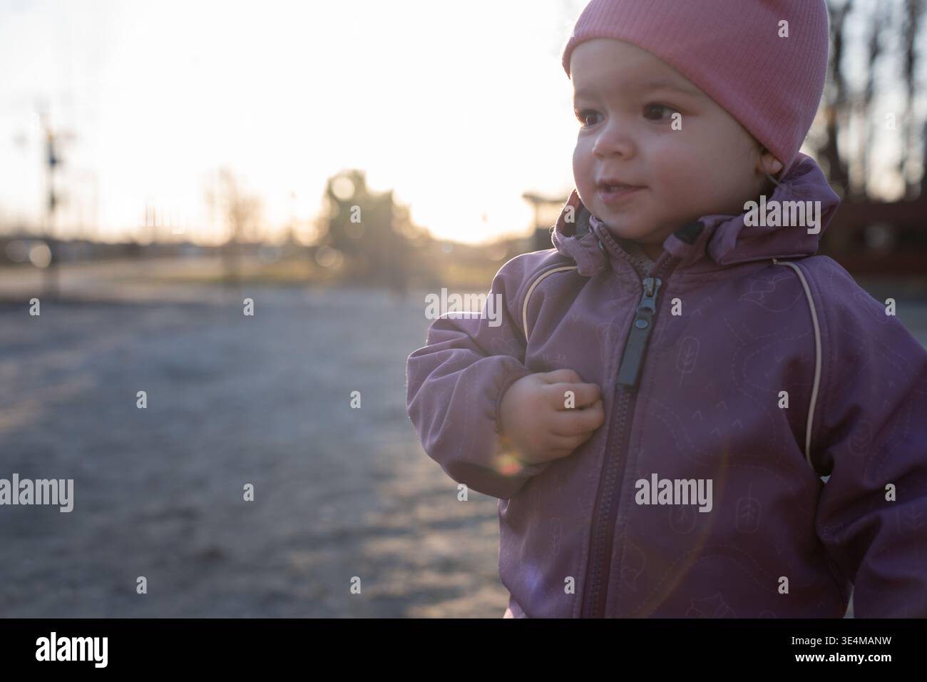 Nahaufnahme eines Kleinkindes in rosa Mütze und malvenfarbenen Jacke, bei Sonnenuntergang draußen mit warmer Hintergrundbeleuchtung, weichem Bokeh und Linsenfleck, gebadet i Stockfoto
