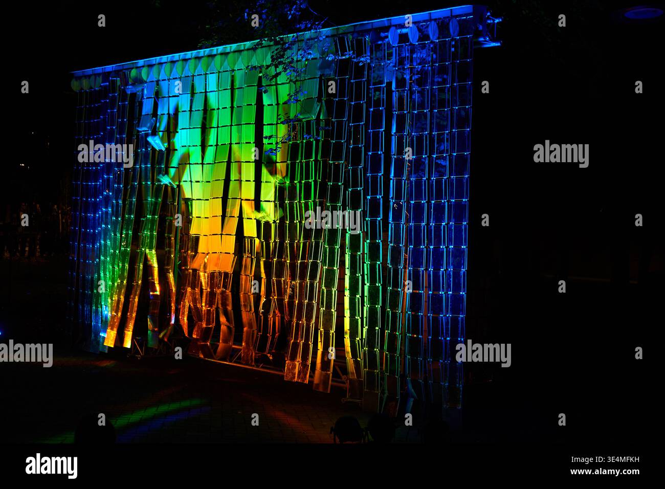 Eine nächtliche Outdoor-Szene mit einer großen Lichtkunstinstallation, bestehend aus hängenden reflektierenden Panels, die in einem Raster angeordnet sind. Die Stockfoto