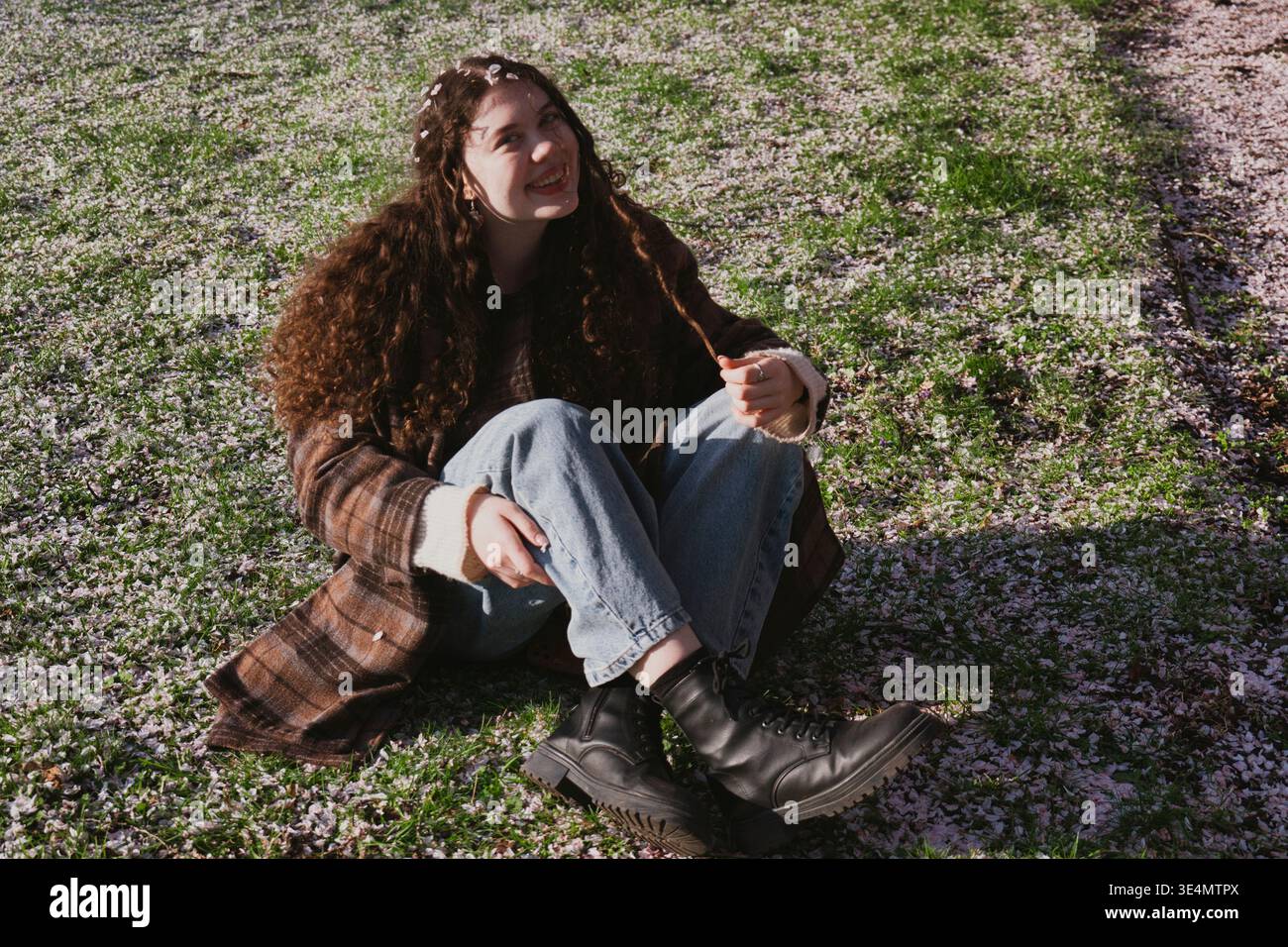 Eine junge Frau, die auf einem mit Kirschblüten bedeckten Gras sitzt. Entspannte Frühlingsszene in einem Park mit sanftem Sonnenlicht. Stockfoto