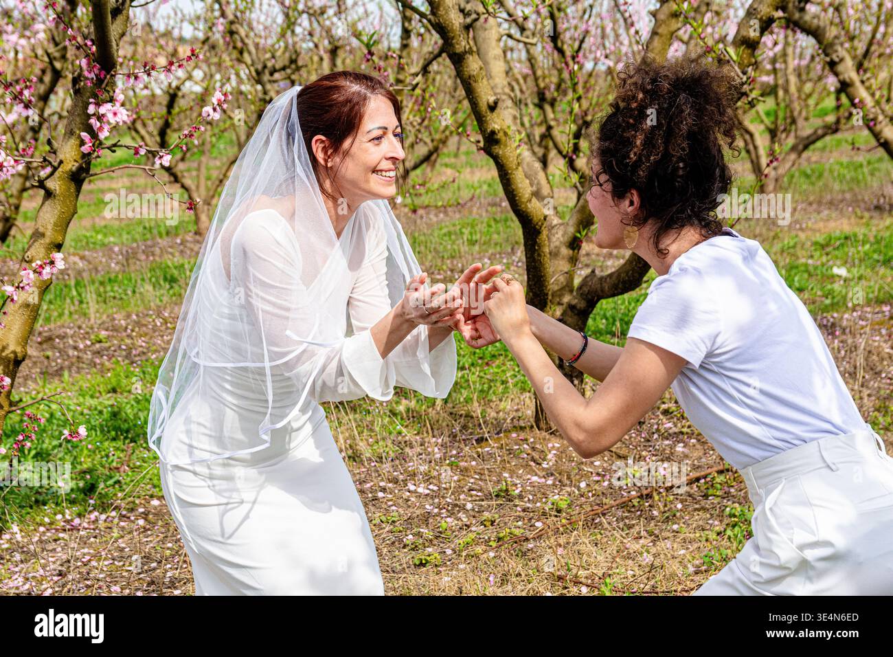Braut in weißem Kleid und Schleier lacht mit fröhlichem Freund zwischen blühenden Frühlingsplantagen, feiert Freundschaft, Liebe und warme offene Momente Stockfoto