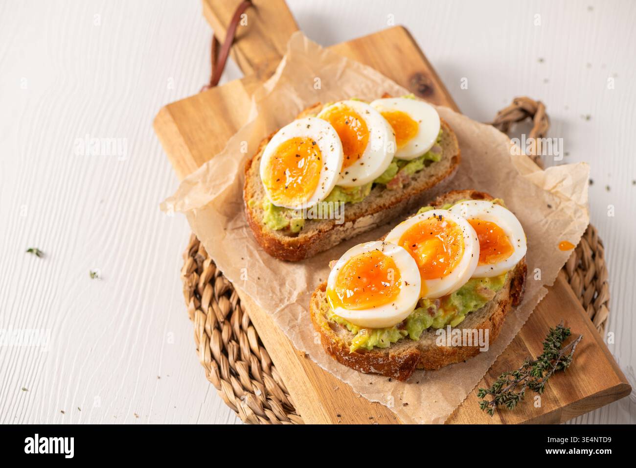 Zwei Scheiben gerösteter Sauerteig, belegt mit Avocadopüree und marmelierten weichen gekochten Eiern. Hochwinkelschuss auf Holzbrett mit Pergamentpapier und Natu Stockfoto