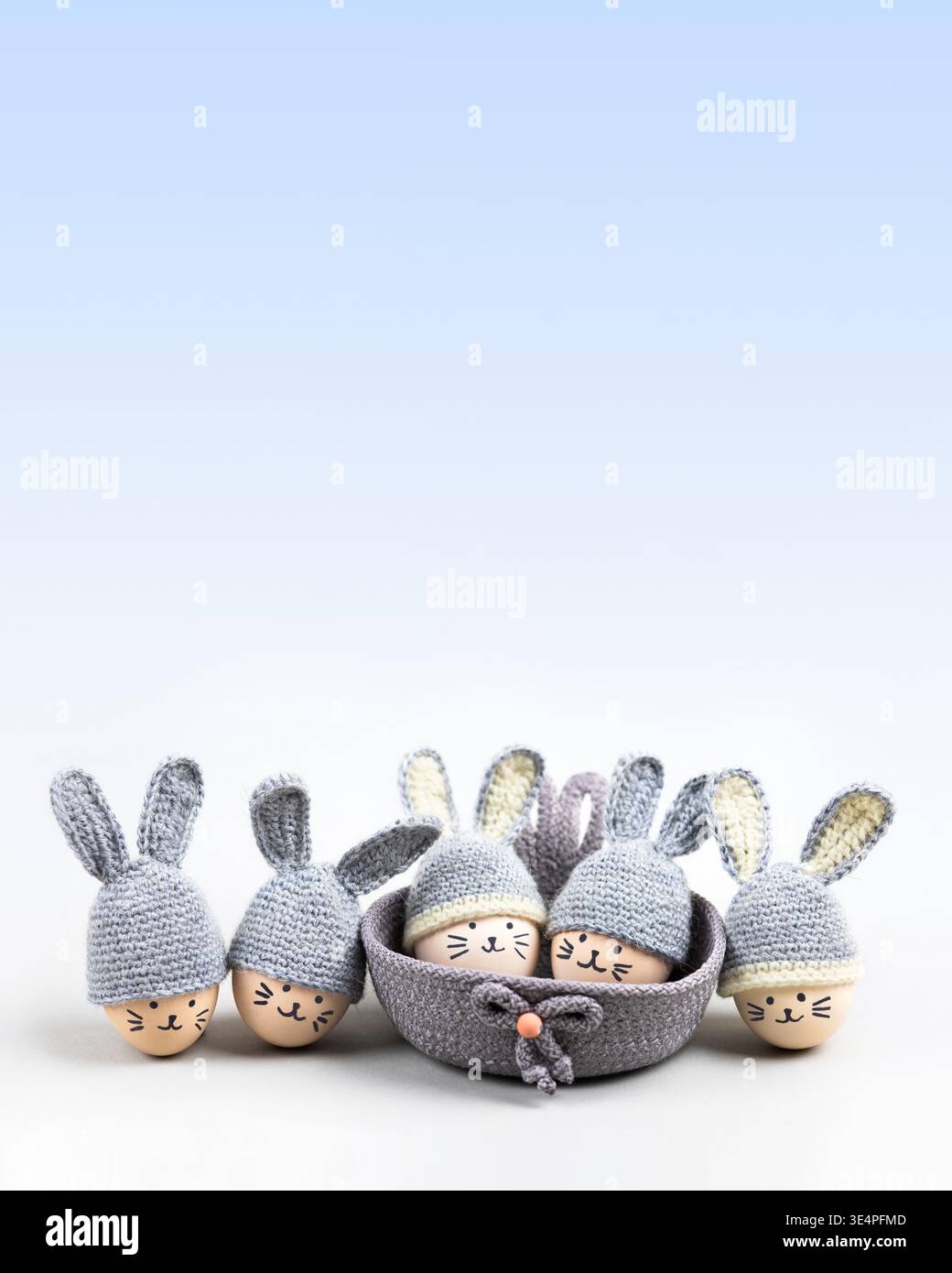 Fünf Ostereier mit lustigen Gesichtern und grauen Häkelhüten mit Hasenohren in einem dekorativen Korb auf einem grauen blauen Hintergrund. Happy Easter-Konzept. Grußkarte. Stockfoto