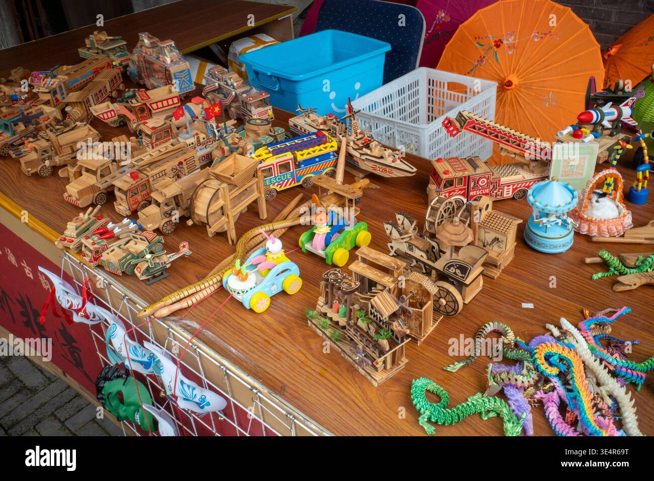 Bunte Holzspielzeuge auf einem Marktstand verstreut. Spielerische Fahrzeuge und Strukturen säumen das Display. Helle Regenschirme und dekorative Gegenstände verleihen Charme Stockfoto
