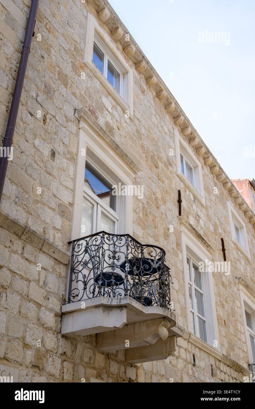 Steinfassade mit weiß gerahmten Fenstern und einem kunstvoll verzierten schmiedeeisernen Balkon, von unten aus vor einem klaren blauen Himmel, mediterrane Geschichte Stockfoto