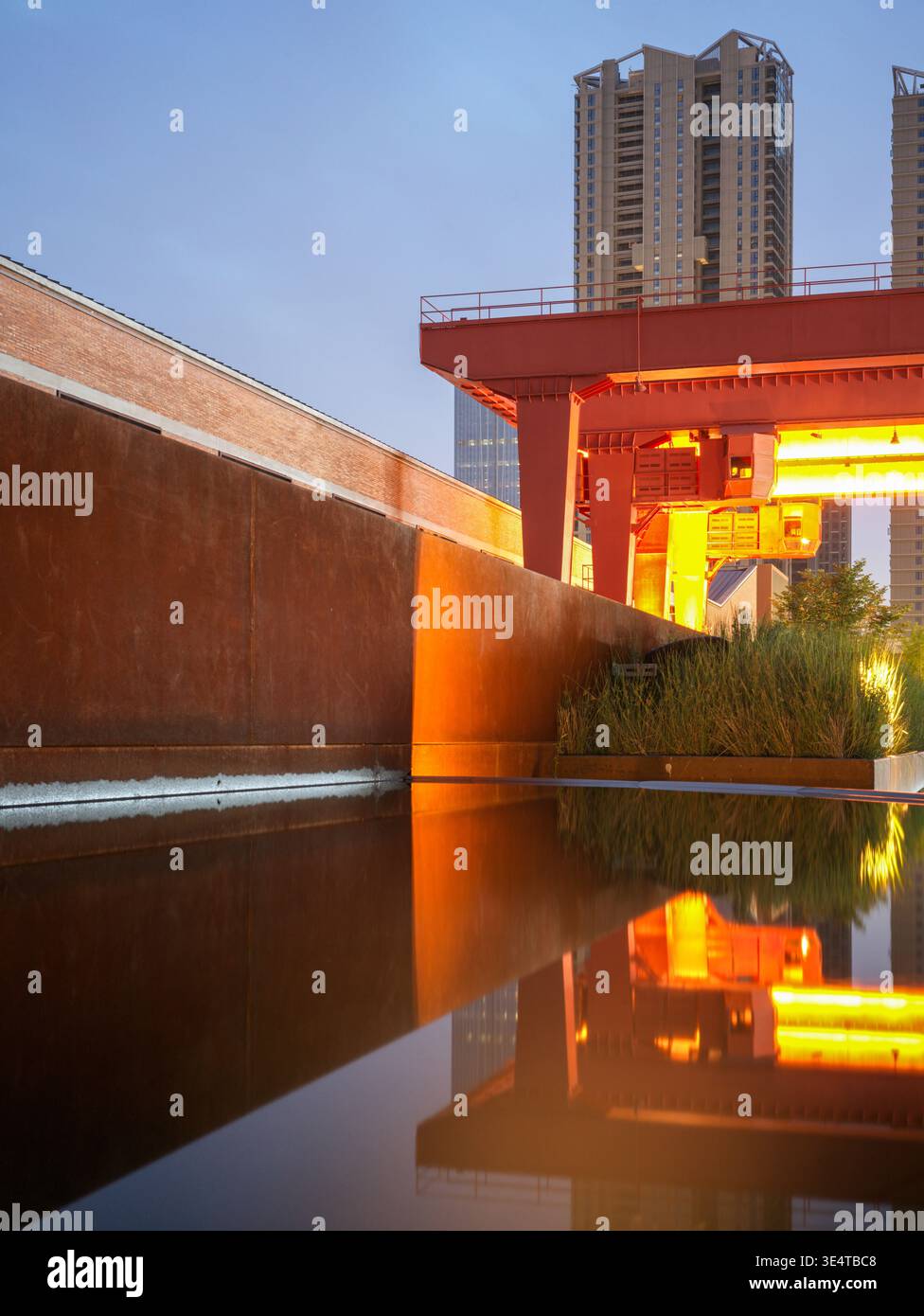Strukturierte Kortenstahlwand, die sich perfekt in einem dunklen Wasserbecken spiegelt, mit einem beleuchteten orangefarbenen Industriekran, der in der Dämmerung dahinter steht. Die sc Stockfoto