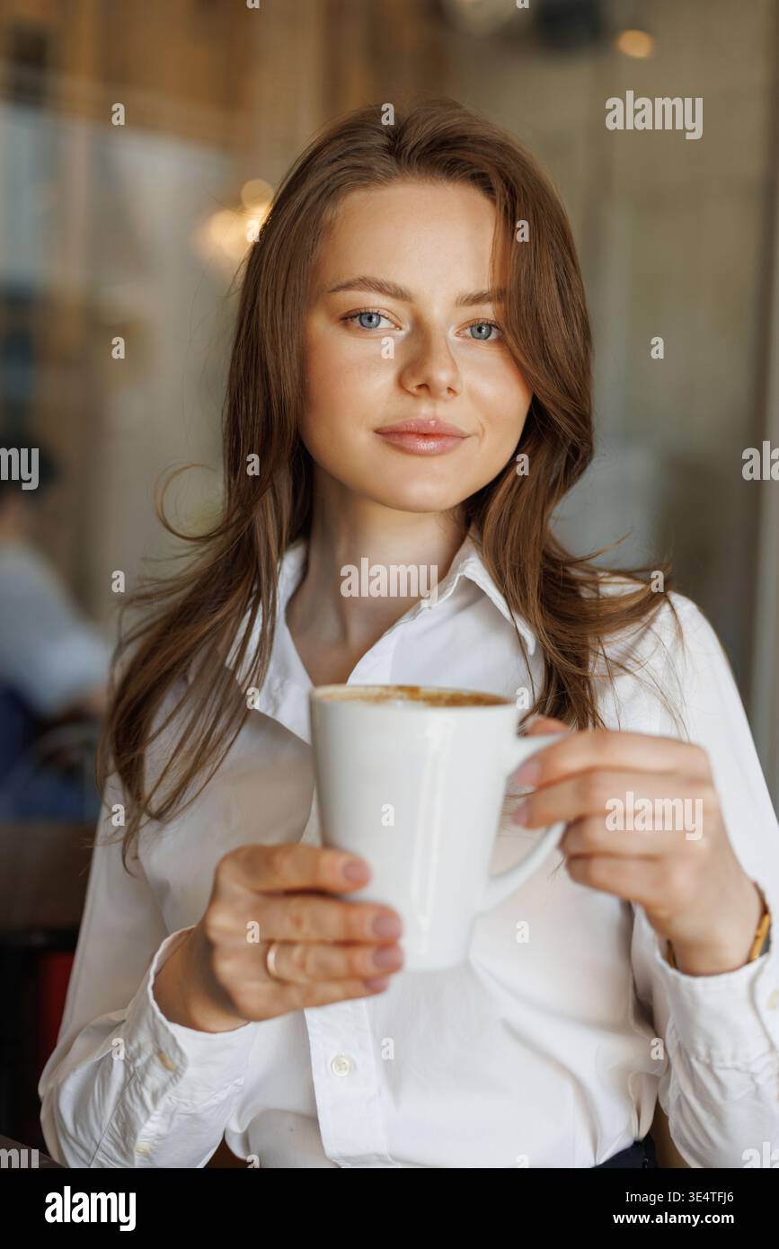 Selbstbewusste Geschäftsfrau, die in einem Café Kaffee trinkt. Eine ungezwungene Arbeitsumgebung, sanftes Licht und freundlicher Ausdruck spiegeln einen entspannten, professionellen Lebensstil wider Stockfoto