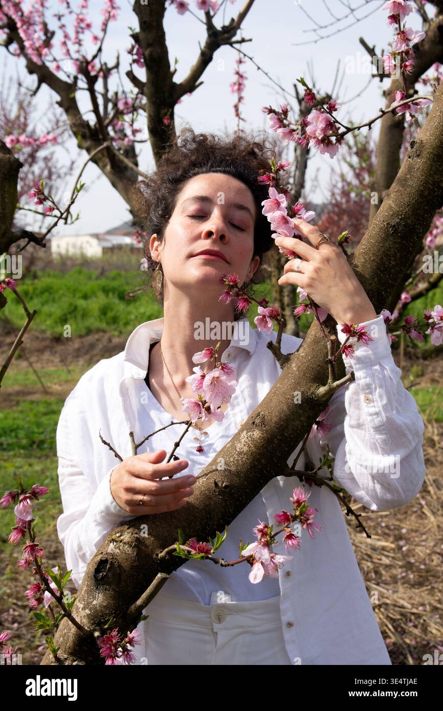 Junge Erwachsene Frau mit geschlossenen Augen, die im Frühling in einem lebhaften Obstgarten, umgeben von zarten Pflanzen, Ruhe findet und sich wieder mit der Natur verbindet Stockfoto