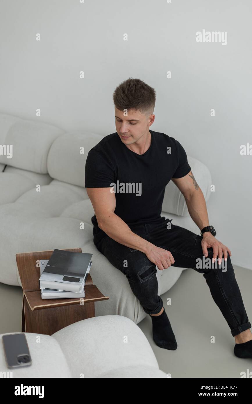 Hübscher erfolgreicher Geschäftsmann in schwarzer Mode, der auf dem Sofa sitzt und zu Hause Zeitschriften hat Stockfoto